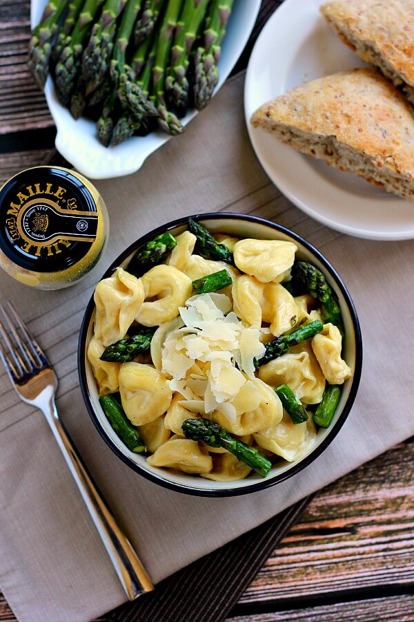 overhead view of tortellini in mustard cream sauce in a bowl next to asparagus, bread, and a jar of mustard. 