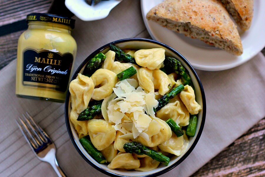 overhead view of tortellini in mustard cream sauce in a bowl next to asparagus, bread, and a jar of mustard. 