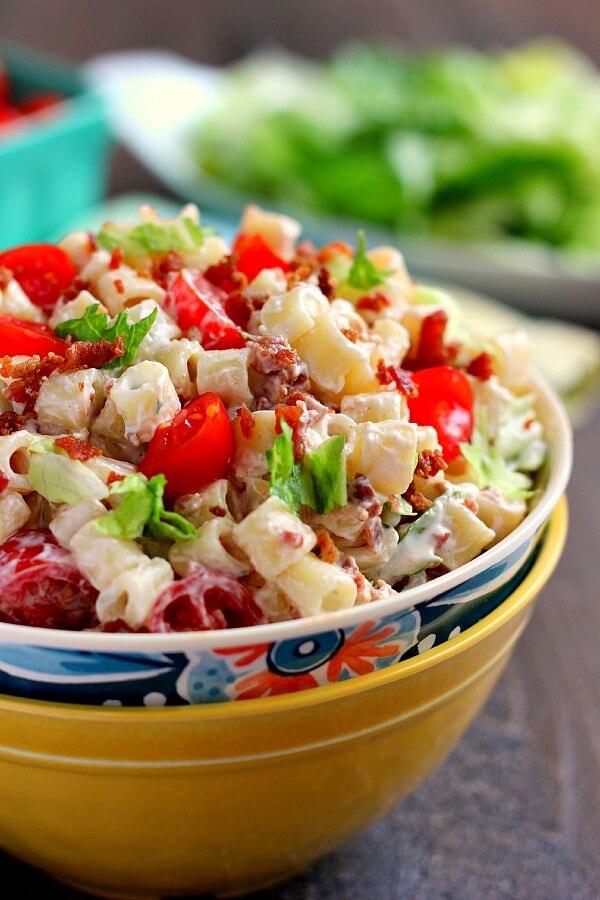 A bowl of BLT pasta salad. Chopped lettuce is blurred in the background. 