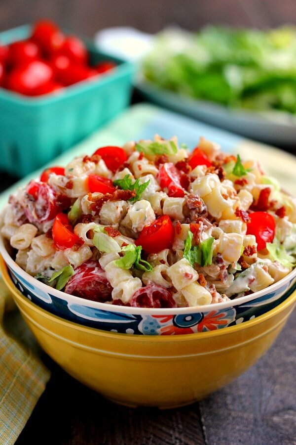 A bowl of easy BLT pasta salad, with cherry tomatoes and lettuce in the background. 