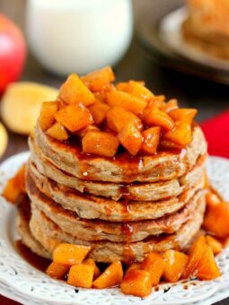 A stack of apple cinnamon pancakes topped with diced cinnamon apples and syrup, served on a white plate, with more apples and a glass of milk in the background.