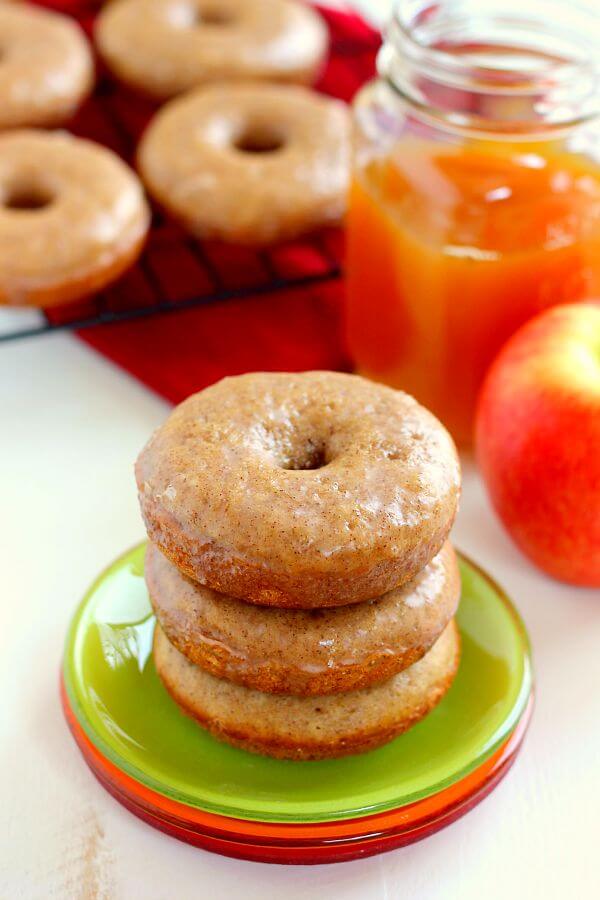 A stack of three baked apple cider donuts. More donuts and a jar of cider rest in the background. 