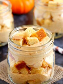 A mason jar filled with layers of creamy no-bake pumpkin cheesecake trifle and cubed cake, set on a burlap cloth with a small pumpkin and more jars blurred in the background.
