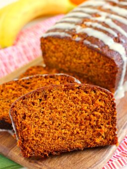 Two thick slices of moist honey glazed pumpkin banana bread rest on a wooden cutting board in front of a loaf drizzled with white icing. A banana and a red-striped cloth are visible in the background.