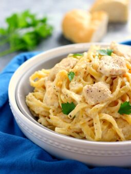 A bowl of creamy chicken fettuccine Alfredo, topped with parsley, sits on a blue cloth. In the background, blurred bread and fresh parsley complete this inviting scene.