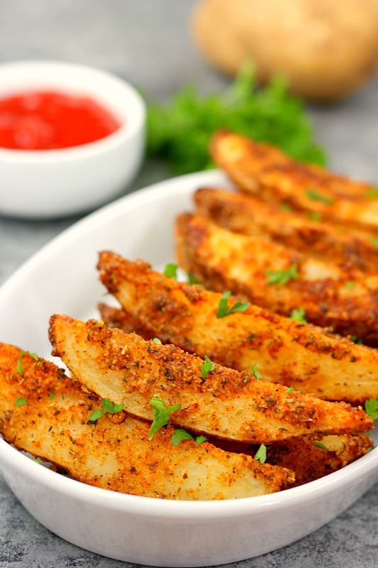 garlic parmesan potato wedges in a white serving dish. A bowl of ketchup is in the background. 