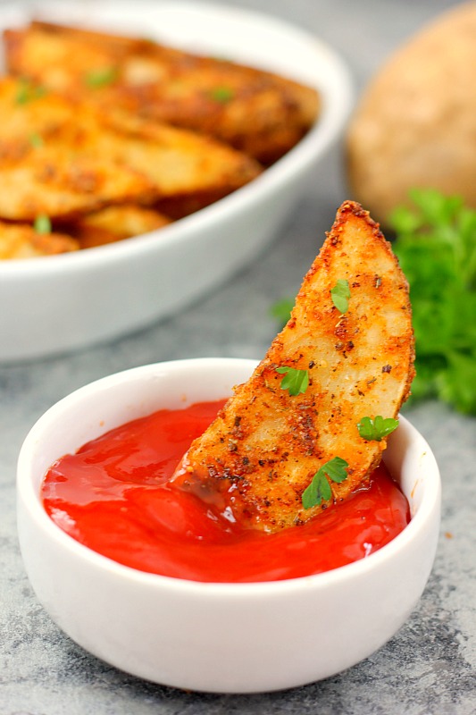 a garlic parmesan potato wedge being dipped into a white bowl of ketchup