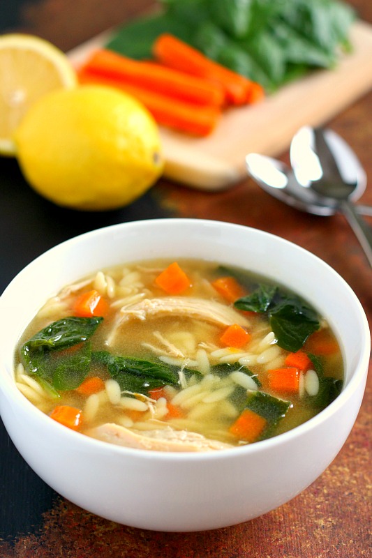 A bowl of lemon chicken orzo soup with lemons, carrots, and spinach on a cutting board in the background. 