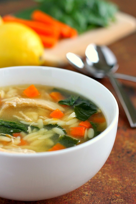 A bowl of lemon chicken orzo soup with lemons, carrots, and spinach on a cutting board in the background. 