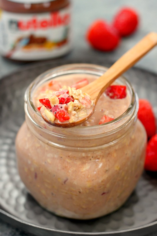 A small wooden spoon resting on a jar filled with Nutella overnight oats with strawberries. 