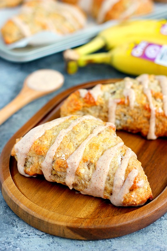 two glazed banana scones on a wood board. bananas and more scones rest in the background. 