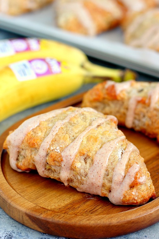 two glazed banana scones on a wood board. bananas and more scones rest in the background. 