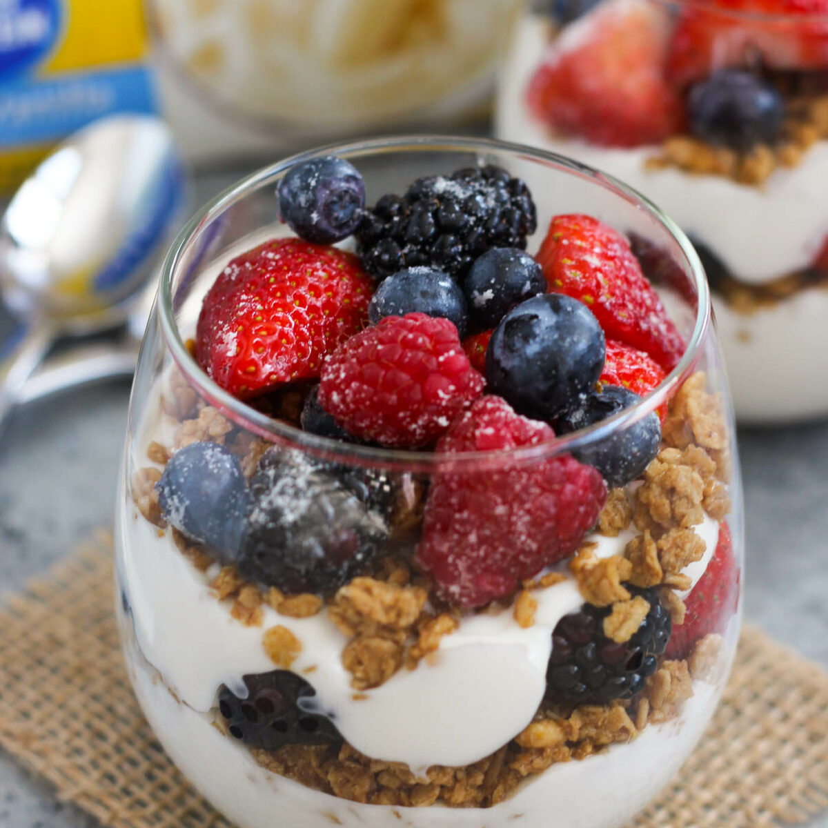 A glass filled with layers of granola and creamy yogurt creates a delicious yogurt parfait recipe, topped with fresh strawberries, raspberries, blueberries, and blackberries, placed on a small burlap mat with a spoon nearby.