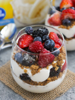 A glass filled with layers of granola and creamy yogurt creates a delicious yogurt parfait recipe, topped with fresh strawberries, raspberries, blueberries, and blackberries, placed on a small burlap mat with a spoon nearby.