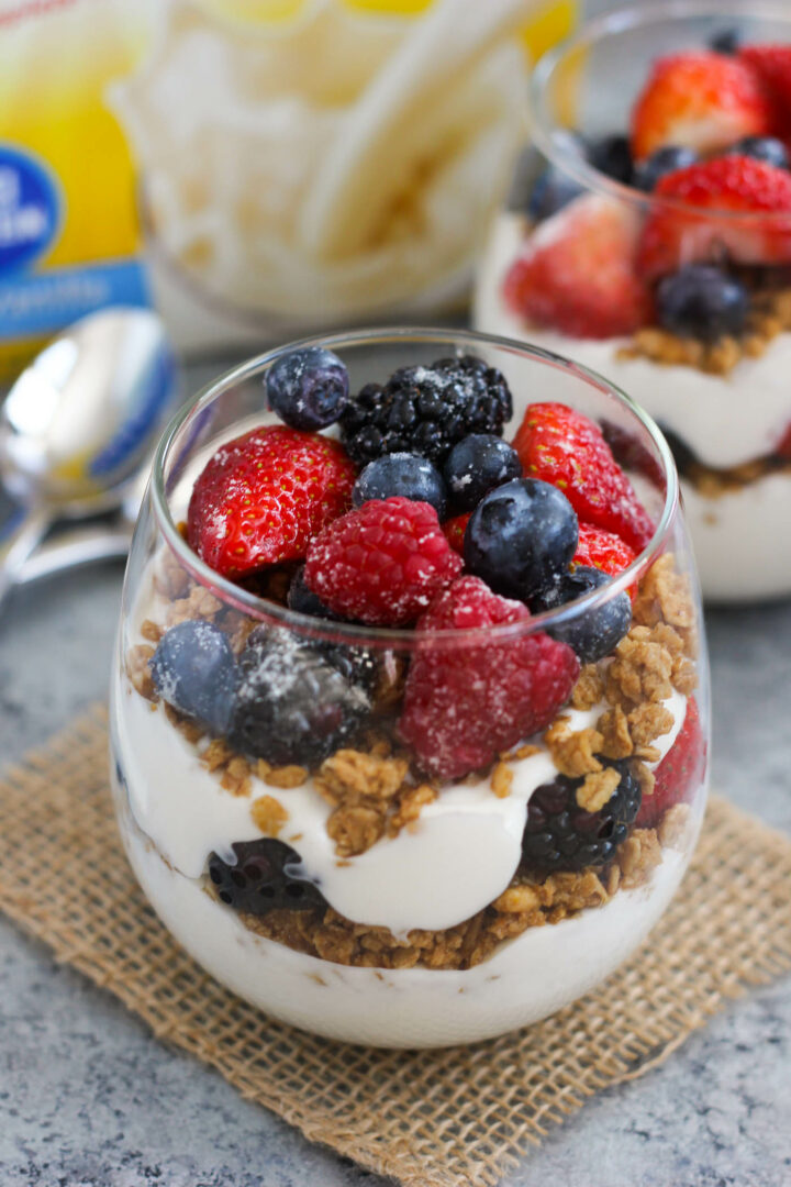 A glass filled with layers of granola and creamy yogurt creates a delicious yogurt parfait recipe, topped with fresh strawberries, raspberries, blueberries, and blackberries, placed on a small burlap mat with a spoon nearby.