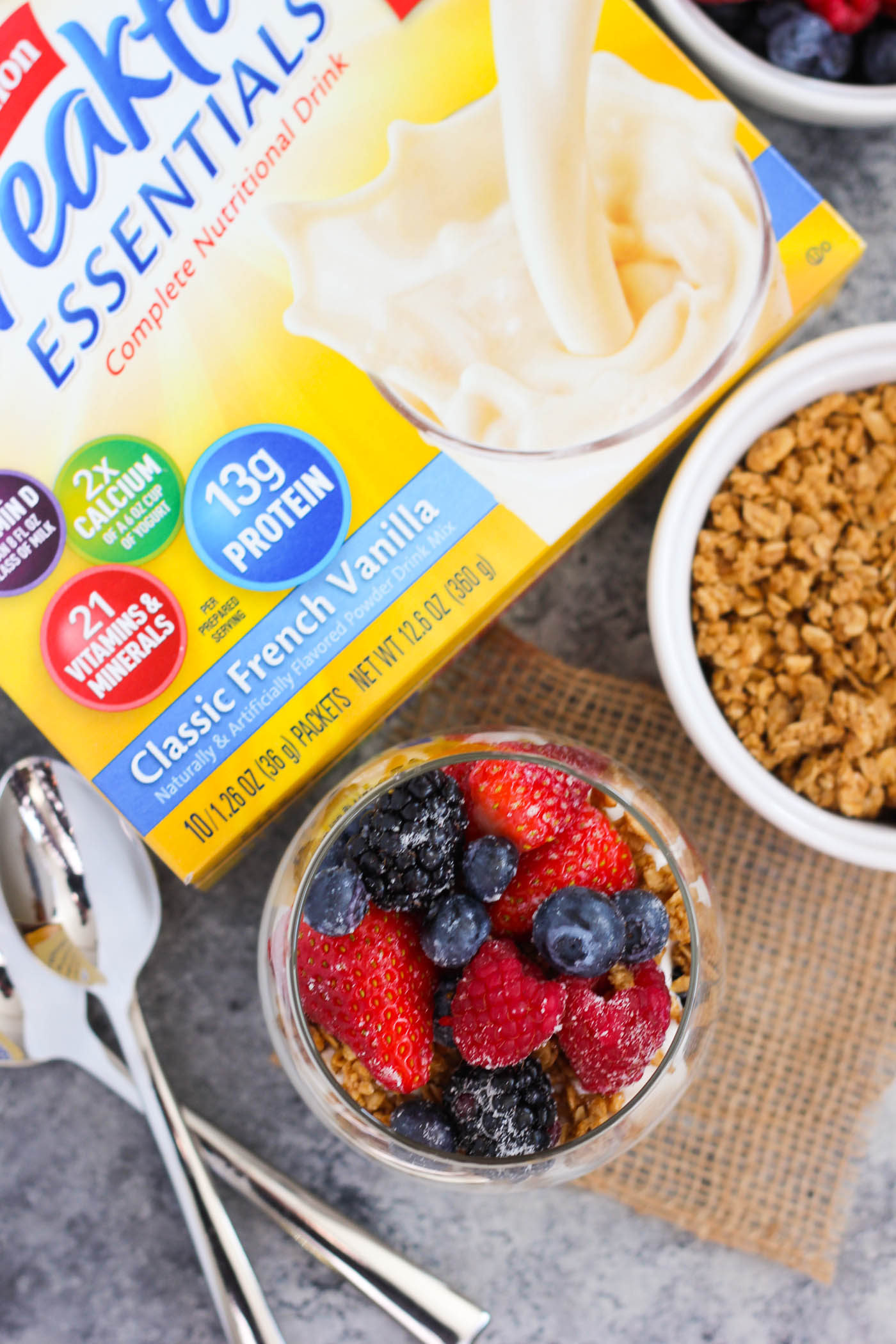 Overhead view of a breakfast parfait, a box of Carnations essential drink powder, and a dish of granola.