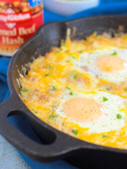 A close-up of a cast iron skillet filled with corned beef hash and eggs, topped with melted cheese and two sunny-side-up eggs, with a can of corned beef hash and a bowl of chopped green onions in the background.