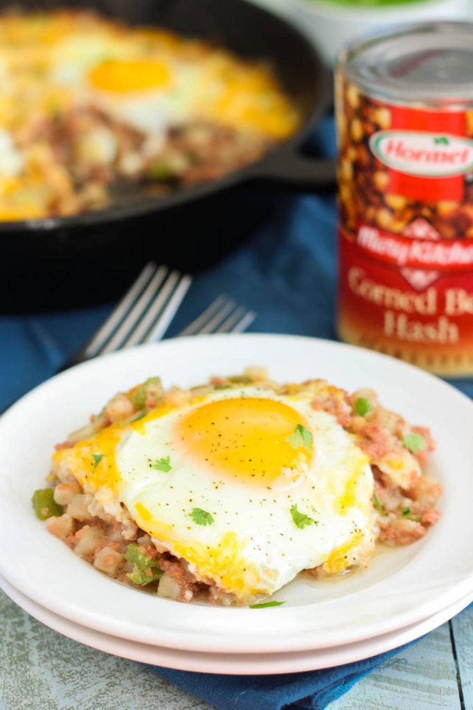 a serving of corned beef and eggs on a small plate. A can of corned beef and a skillet of corned beef hash rest in the background. 