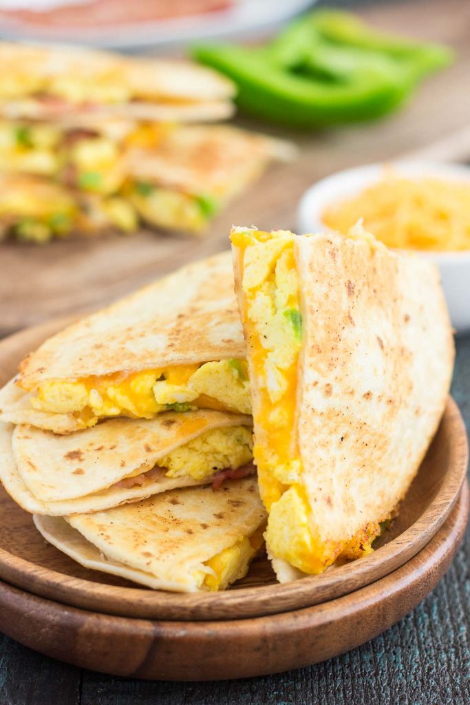 A plate with sliced breakfast quesadillas filled with scrambled eggs, melted cheese, and green peppers. In the blurred background, more breakfast quesadilla slices and fresh green pepper are visible.