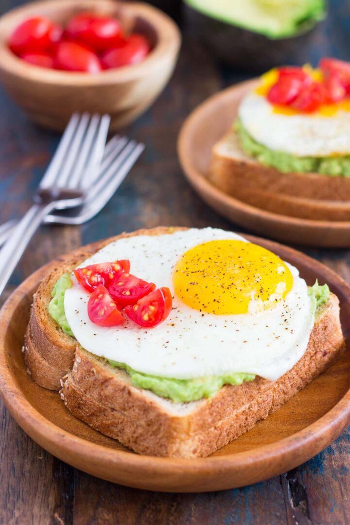 A slice of toasted bread topped with mashed avocado, a fried egg, and sliced cherry tomatoes, served on a wooden plate with another plate of fried egg and avocado toast and bowls of tomatoes in the background.