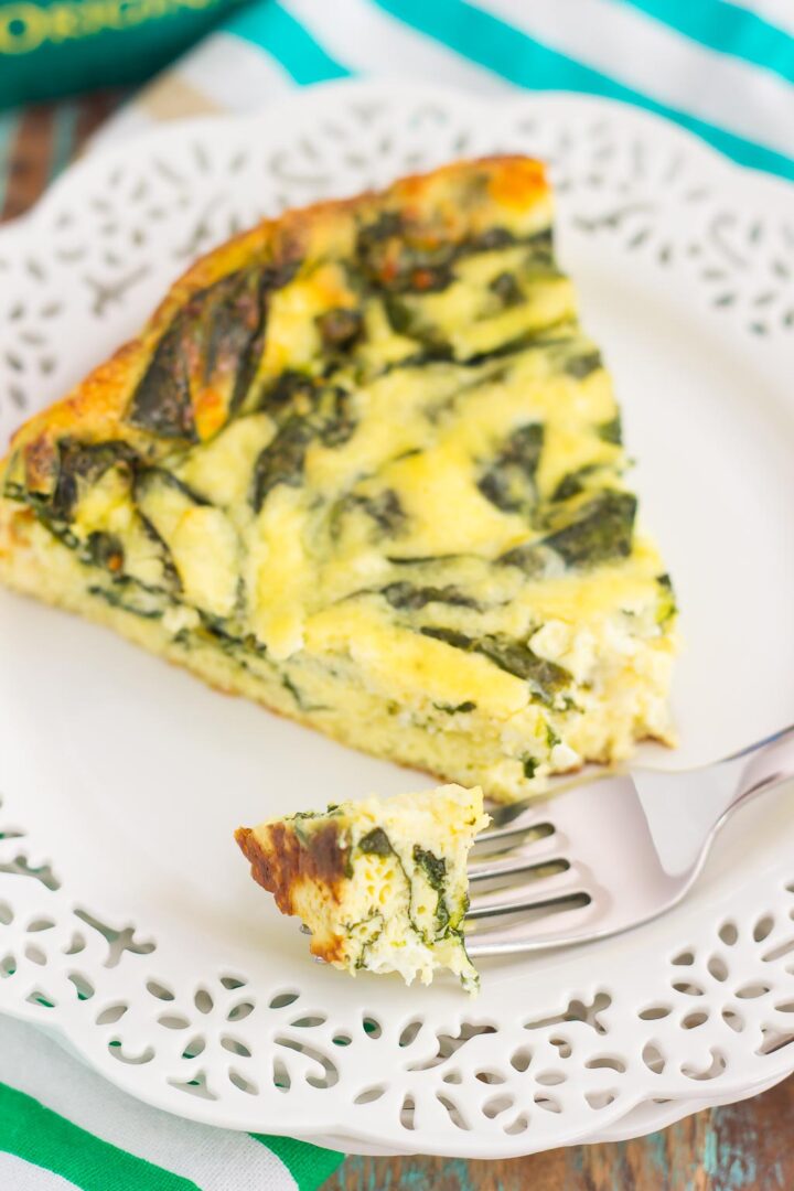 A slice of spinach and cheese frittata, reminiscent of a spinach and feta frittata, sits on a decorative white plate, with a fork holding a small bite, atop a green and white striped cloth.