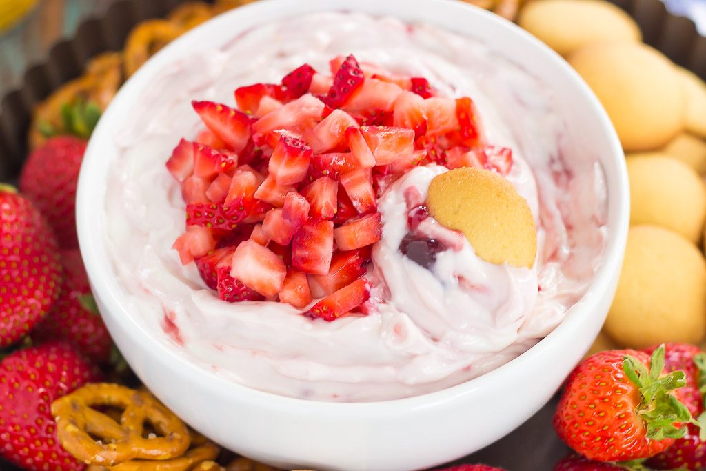 A bowl of strawberry fruit dip topped with fresh strawberries and surrounded by dippers. 