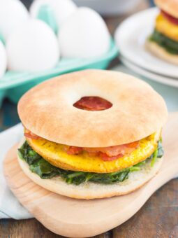 A breakfast sandwich with eggs, sautéed spinach, and tomato on a bagel thin, served on a wooden board. In the background, another breakfast sandwich and whole eggs are visible.