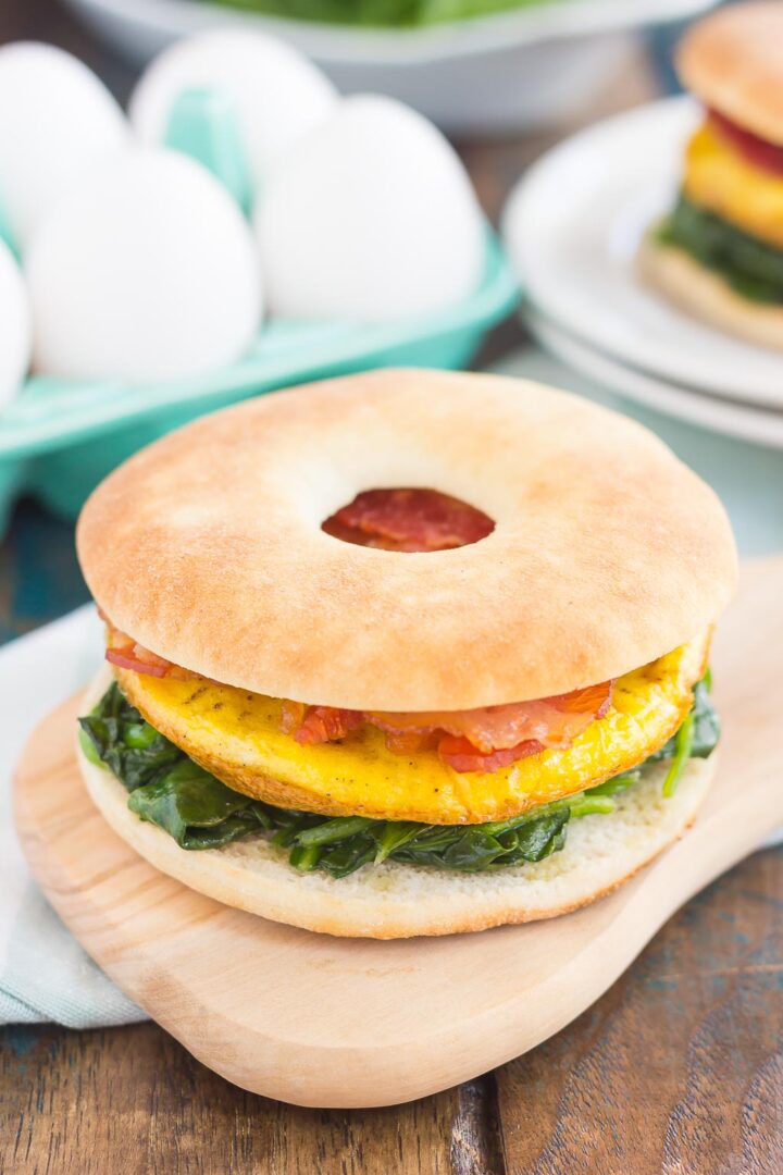 A breakfast sandwich with eggs, sautéed spinach, and tomato on a bagel thin, served on a wooden board. In the background, another breakfast sandwich and whole eggs are visible.