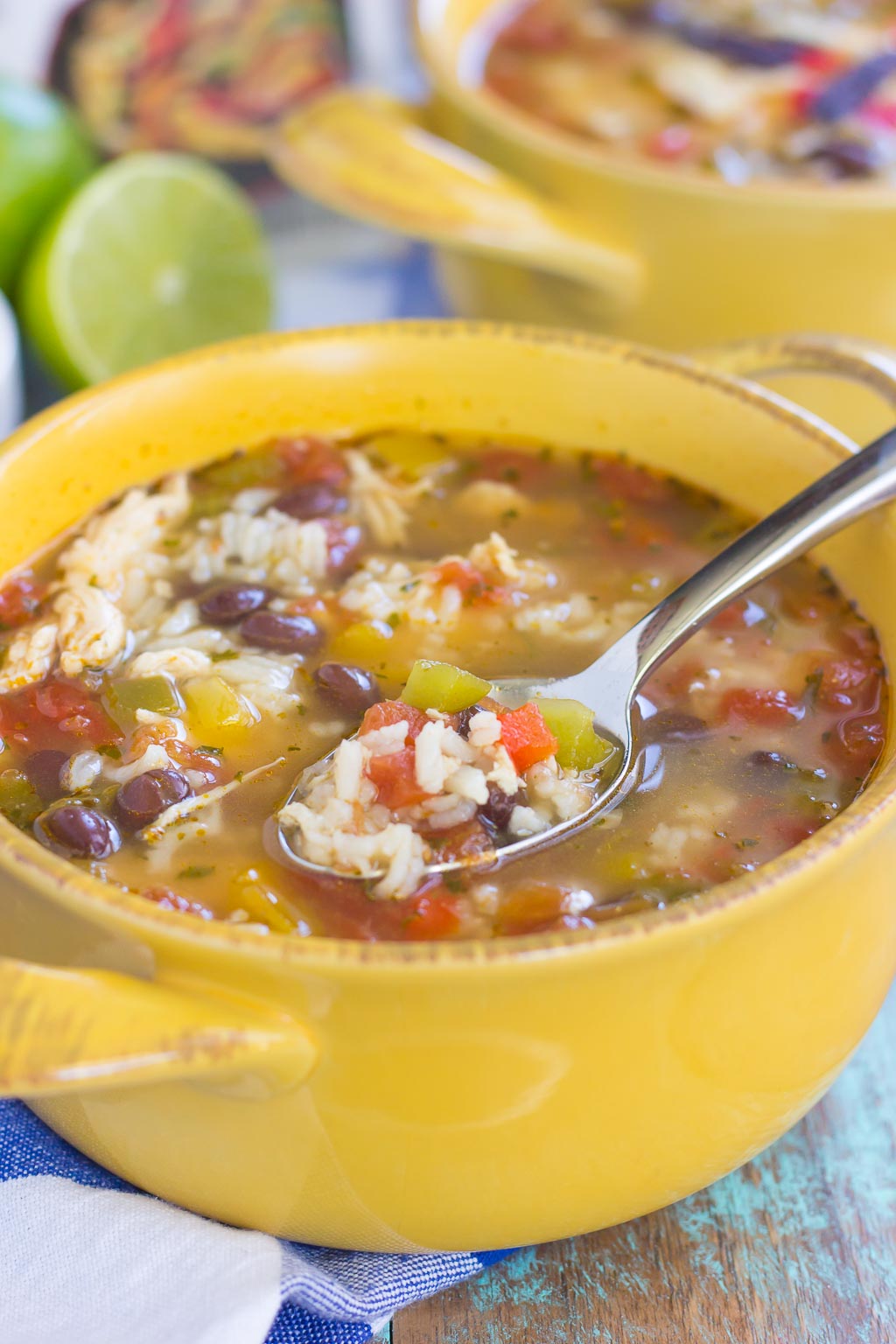 A yellow bowl filled with chicken fajita soup—packed with rice, beans, and vegetables. A spoon rests on the bowl, holding a flavorful bite. Another bowl and lime wedges are visible in the background.