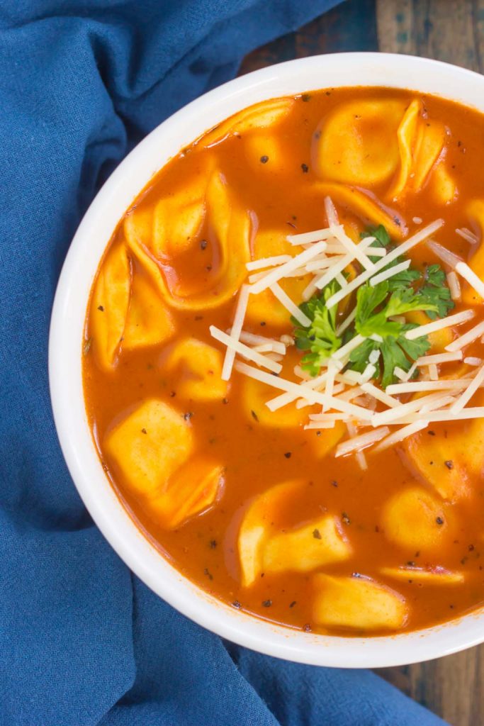 overhead view of tomato tortellini soup garnished with cheese and parsley in a white bowl. The bowl rests on a blue tea towel. 