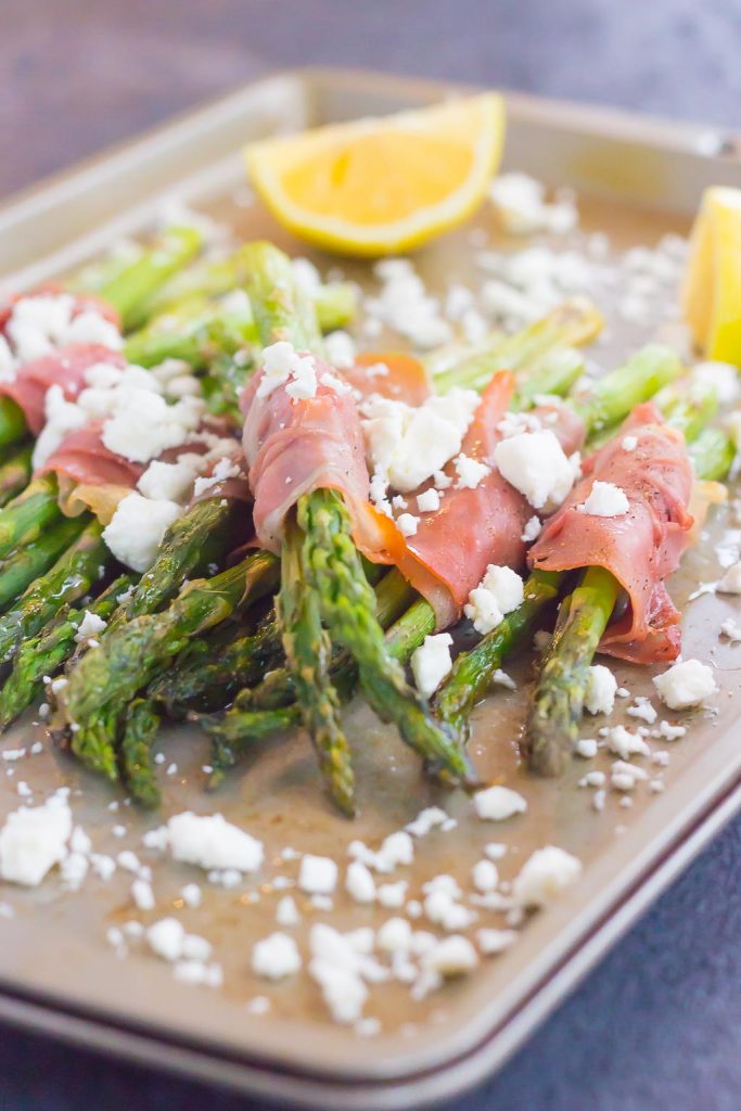 prosciutto wrapped asparagus bundles sprinkled with feta on a baking sheet