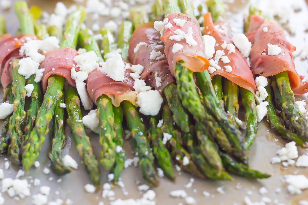 prosciutto wrapped asparagus bundles sprinkled with feta on a baking sheet