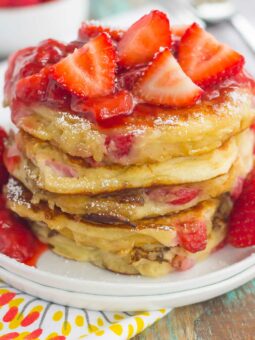 A stack of fluffy strawberry pancakes topped with sliced strawberries and strawberry sauce sits on a white plate, with a colorful napkin and fresh strawberries in the background.