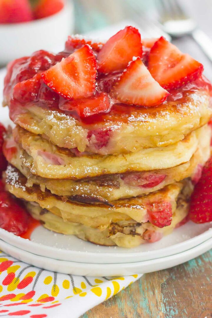 A stack of fluffy strawberry pancakes topped with sliced strawberries and strawberry sauce sits on a white plate, with a colorful napkin and fresh strawberries in the background.