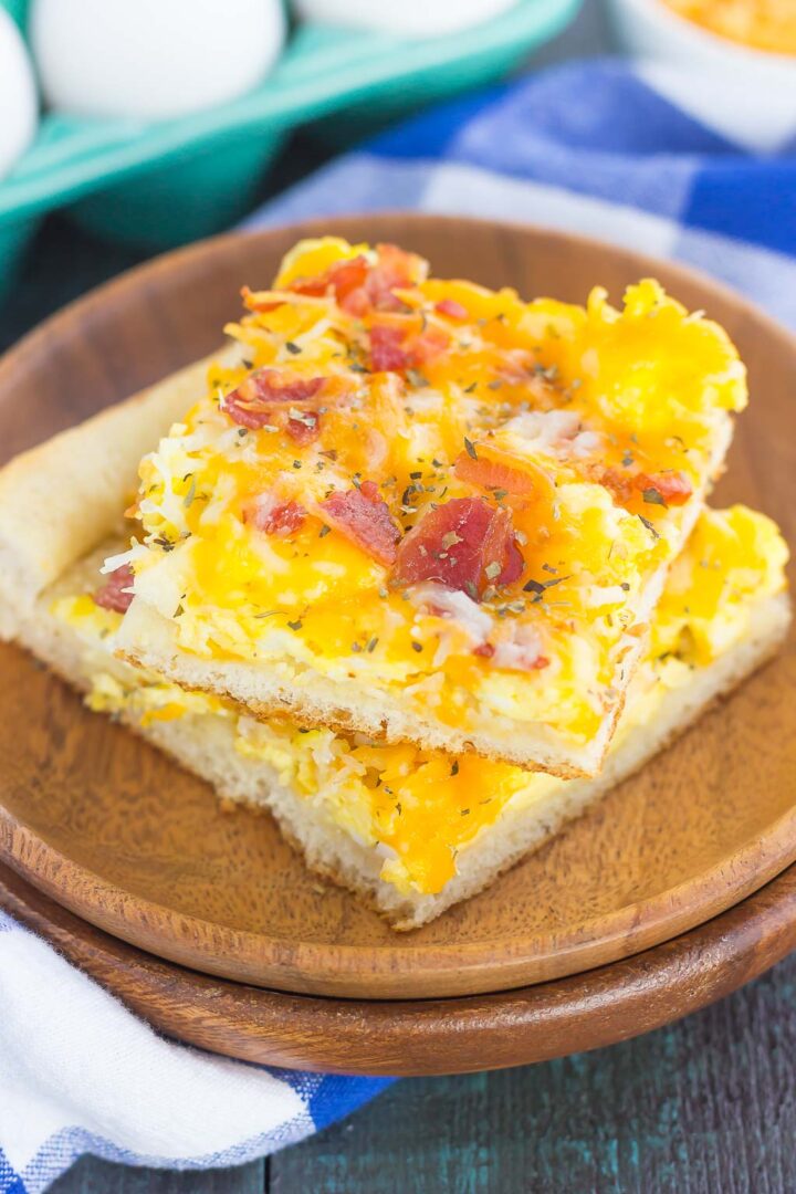 Two square slices of breakfast pizza recipe with eggs, topped with scrambled eggs, melted cheddar cheese, bacon bits, and herbs, served on a wooden plate with a blue and white checkered napkin nearby.