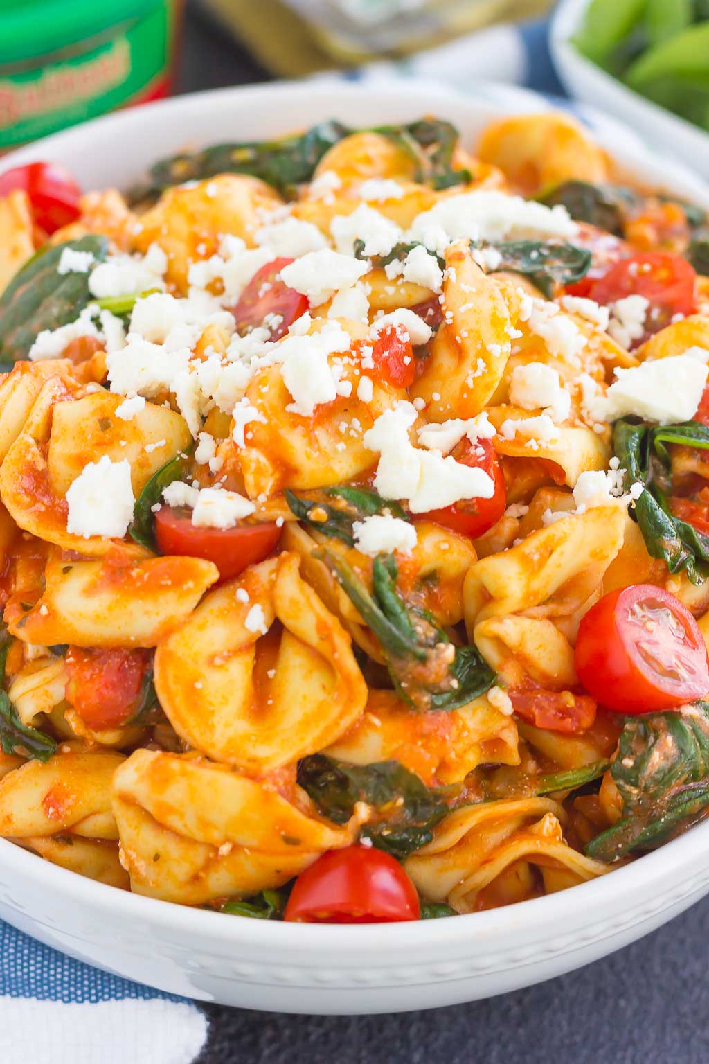 A bowl of spinach and feta tortellini mixed with cherry tomatoes and fresh spinach in a tomato-based sauce, topped with crumbled white cheese.