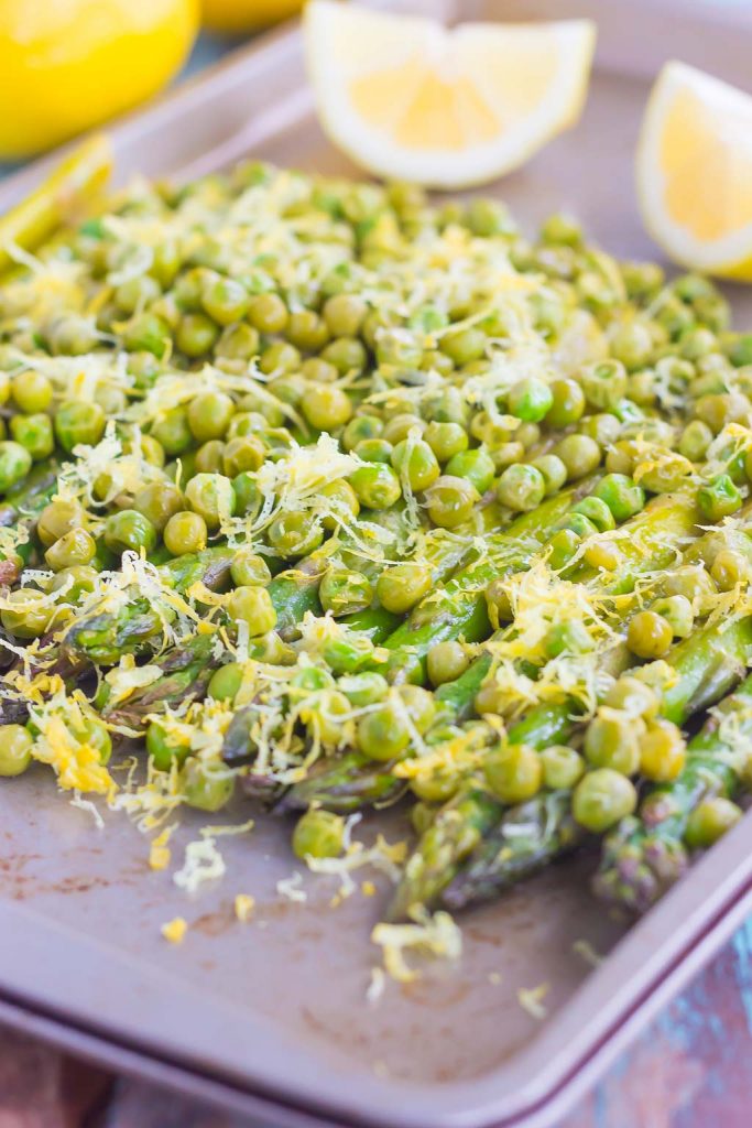 roasted asparagus with lemon and peas on a baking sheet. Lemon wedges rest in the background. 