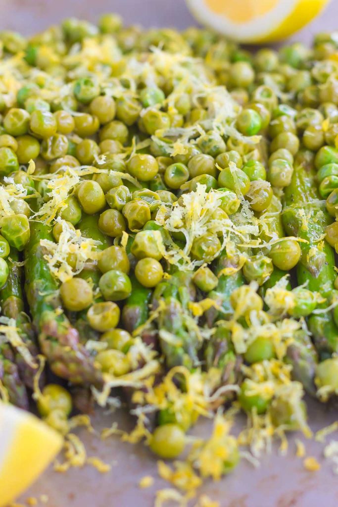 roasted asparagus with lemon and peas on a baking sheet. Lemon wedges rest in the background. 