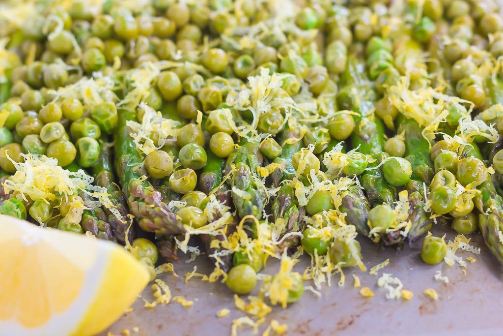 Lemon asparagus and peas on a baking sheet. A lemon wedge rests in the foreground. 