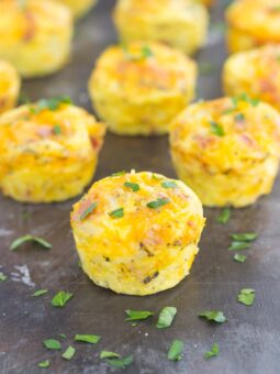 Mini hash brown egg cups topped with melted cheese and chopped herbs are arranged on a dark surface, with one muffin in the foreground and others blurred in the background.