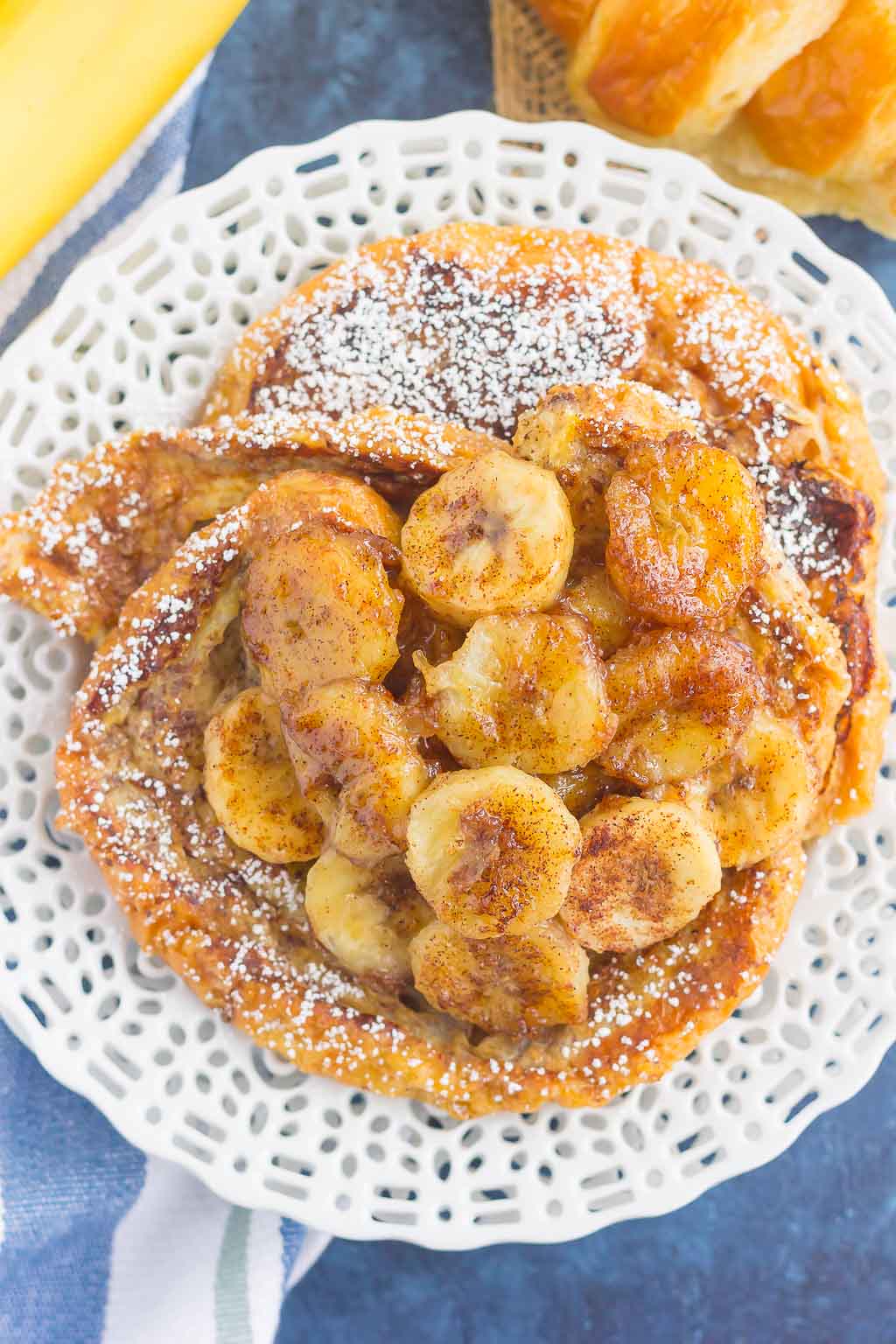 A plate of croissant French toast topped with caramelized banana slices and sprinkled with powdered sugar, served on a white decorative plate.
