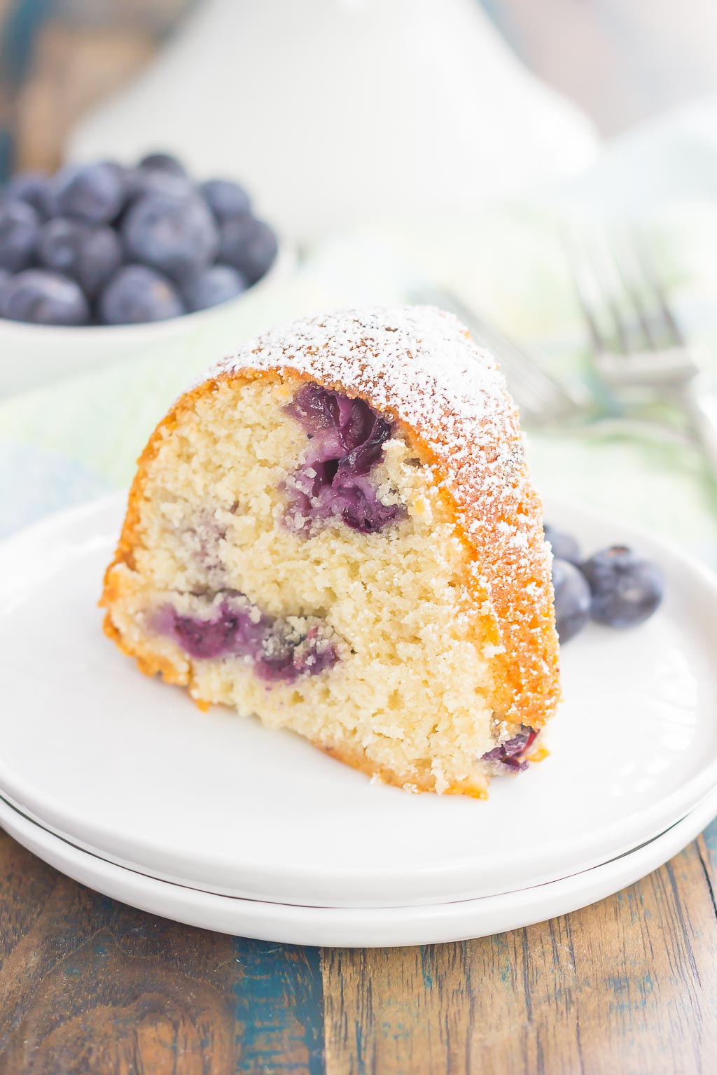 A slice of blueberry yogurt cake on a white plate. A dish of blueberries rest in the background. 