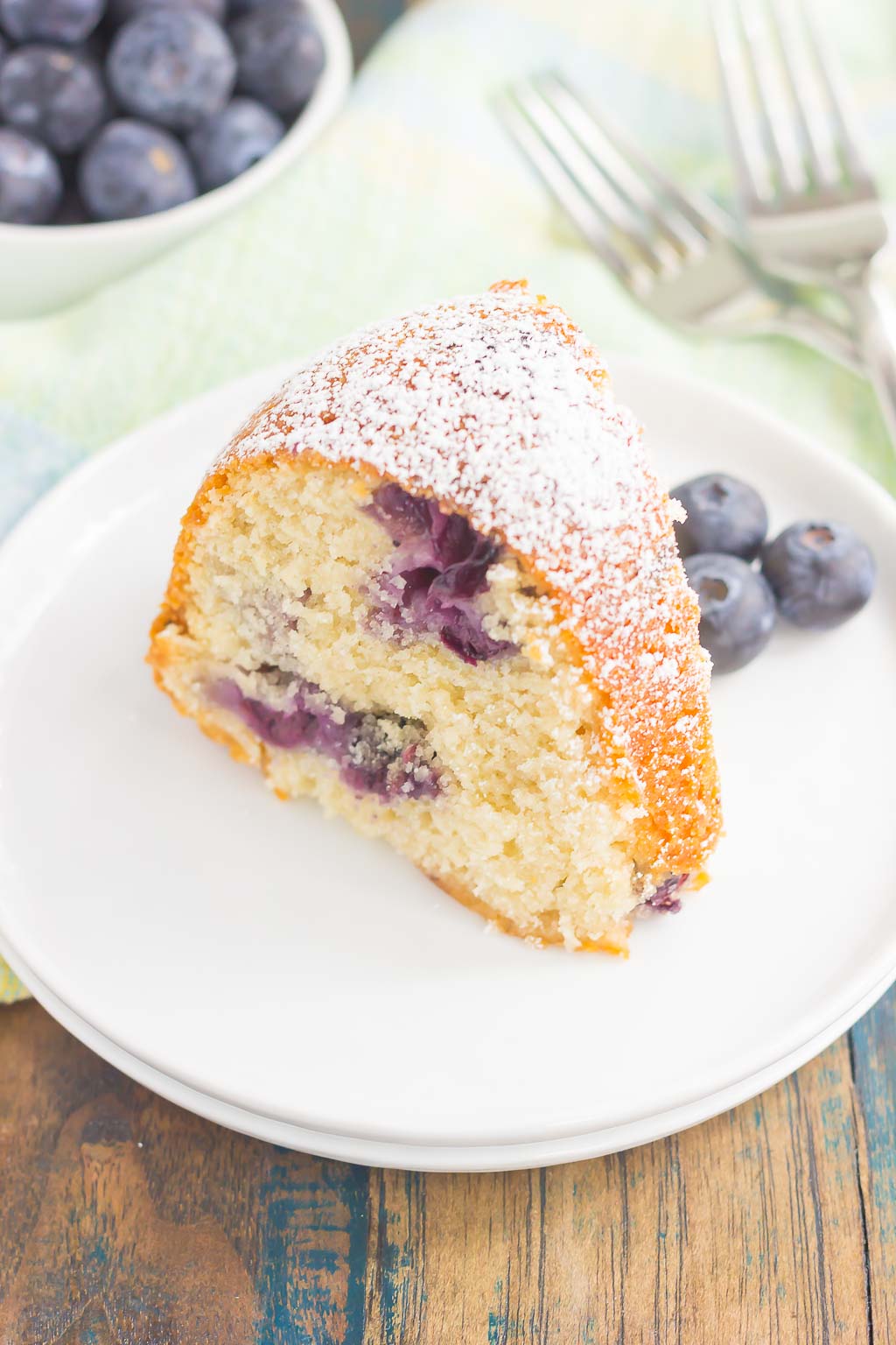 A slice of blueberry bundt cake dusted with powdered sugar on a white dessert plate. A dish of fresh berries rests in the background. 