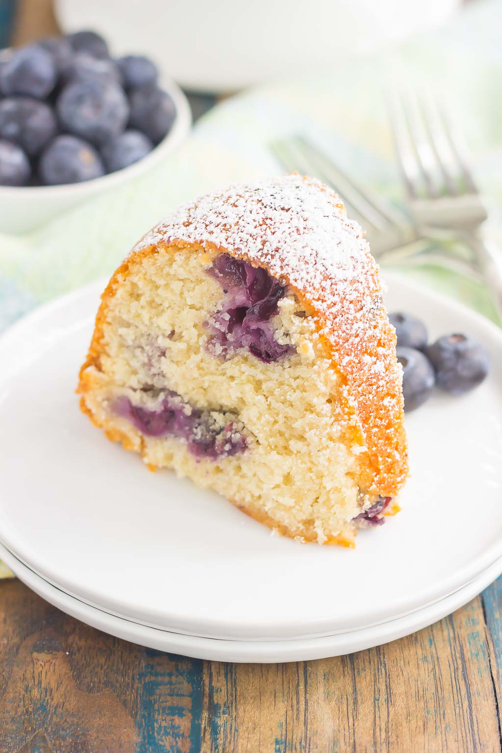 A slice of blueberry bundt cake dusted with powdered sugar on a white dessert plate. A dish of fresh berries rests in the background. 