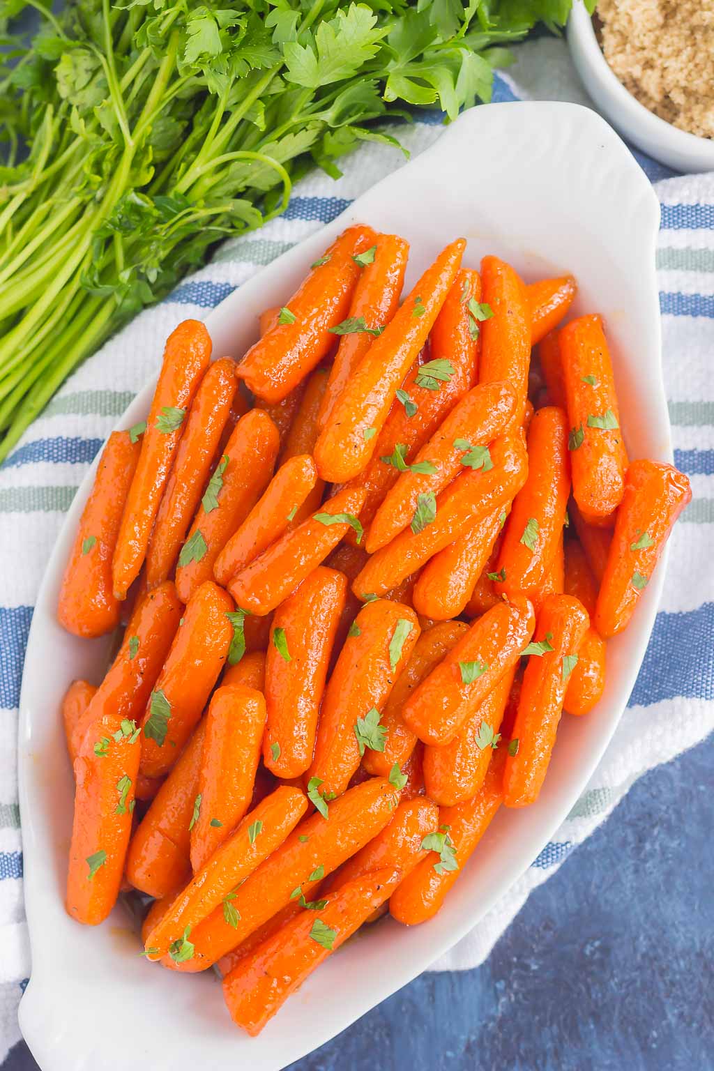 dish of brown sugar glazed carrots atop a tea towel. A bunch of parsley rests in the background. 