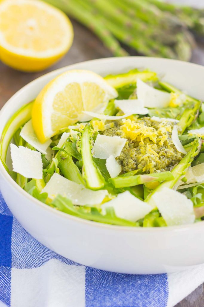 A large white bowl of shaved asparagus salad. Fresh asparagus and a lemon half rest in the background.
