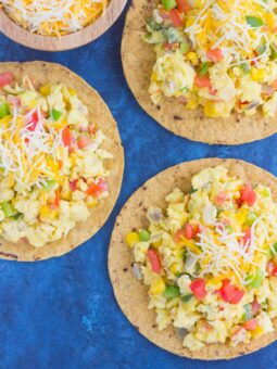 Three southwest scramble tostadas topped with scrambled eggs, chopped vegetables, corn, diced tomatoes, and shredded cheese are arranged on a blue surface. A wooden bowl of extra shredded cheese sits nearby.