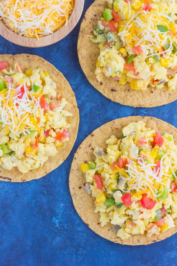 Three southwest scramble tostadas topped with scrambled eggs, chopped vegetables, corn, diced tomatoes, and shredded cheese are arranged on a blue surface. A wooden bowl of extra shredded cheese sits nearby.