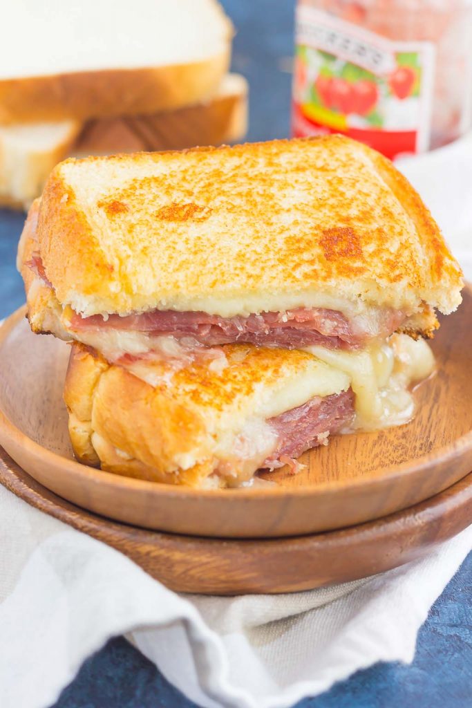 A prosciutto grilled cheese sandwich cut in half, with melted cheese oozing out, stacked on a wooden plate. Slices of bread and a jar of raspberry jam are blurred in the background.
