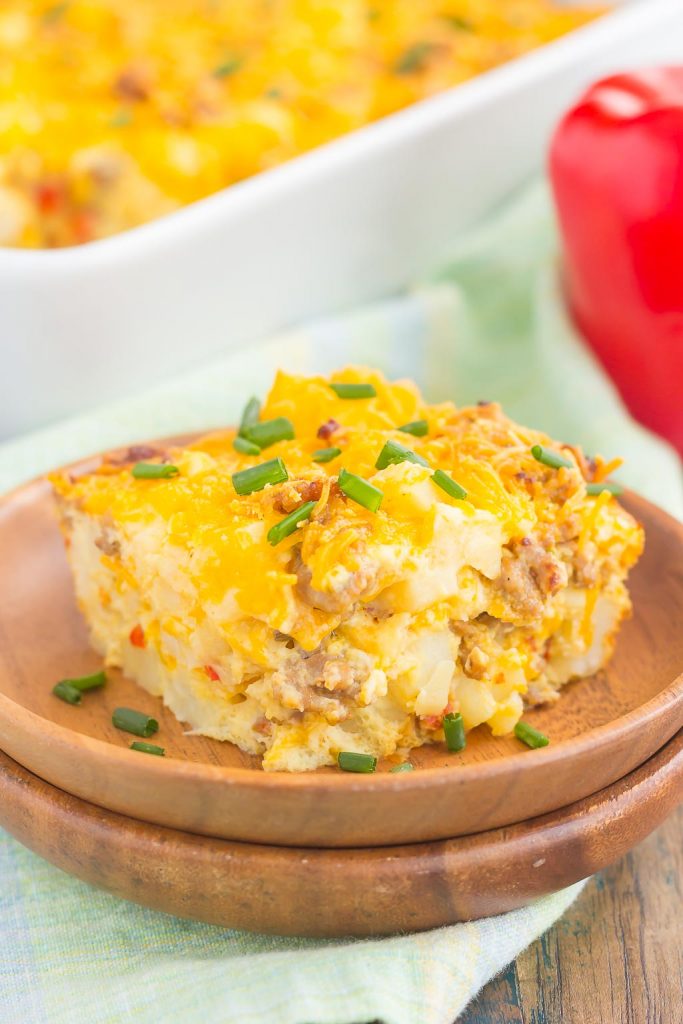 A slice of cheesy sausage hashbrown breakfast casserole with eggs, potatoes, and chopped chives on top, served on a wooden plate with a red bell pepper and casserole dish in the background.